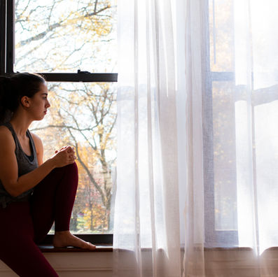 A woman in a gray tank top and red leggings sits by a window, gazing outside. Sunlight filters through sheer curtains, creating a peaceful mood.