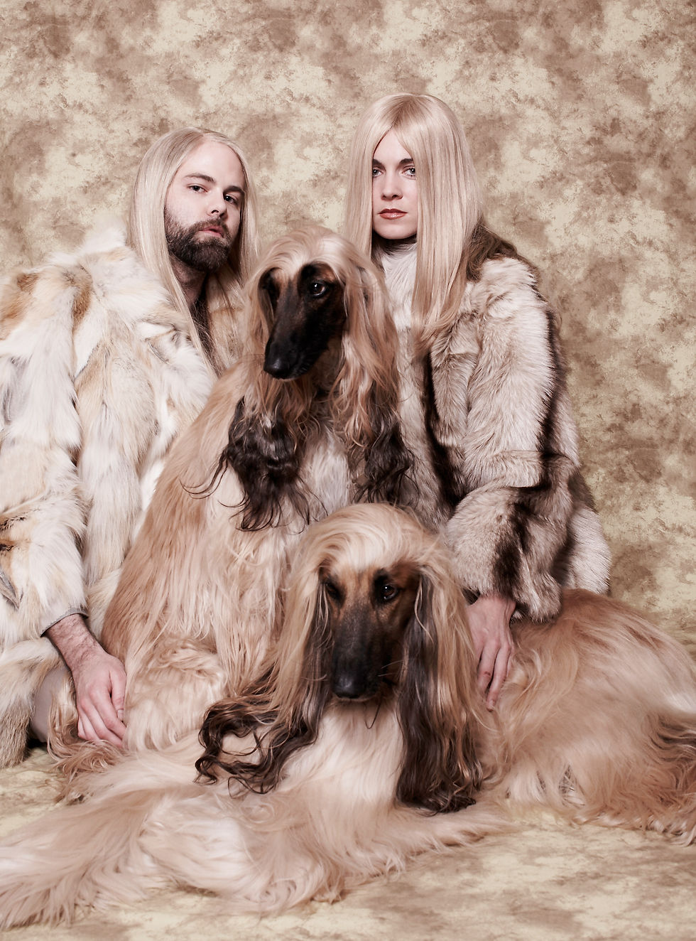 Two people in fur coats, each with long blonde hair, sit with two Afghan Hounds on a beige textured background, creating a harmonious look.