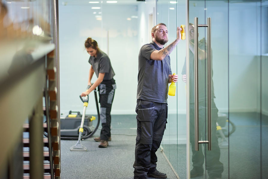 A man cleans a glass door with a cloth, holding a spray bottle, while a woman vacuums the carpet in an office