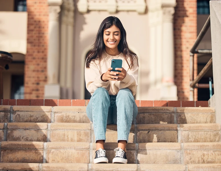 Young woman smiling, using her phone, sits on steps outdoors. EnnovateX IT
