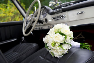 A white wedding bouquet is placed on a black leather seat inside a vintage car
