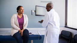 Smiling patient sits on exam table as doctor in white coat talks in a bright clinic room beside a monitor.