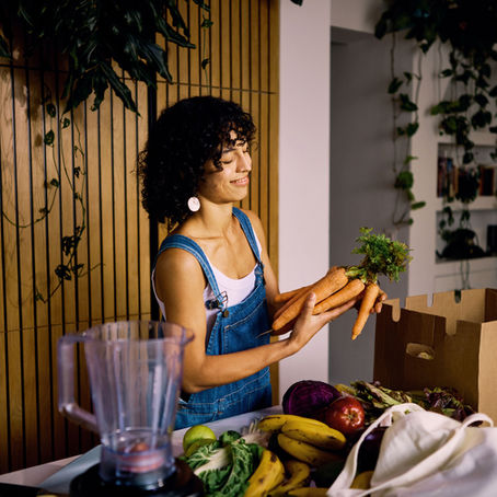 Woman in denim overalls holds carrots, smiling in a kitchen. Surrounded by bananas, cabbage, cardboard box. Plants in the background.