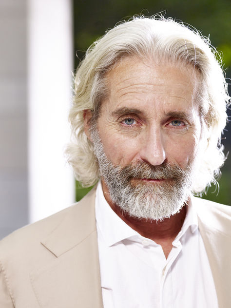 Homem de meia-idade com cabelo e barba grisalhos, vestido com blazer claro, olhando serenamente para a câmara.