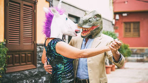 People wearing a unicorn and dinosaur masks dance in a courtyard. One wears a sequin dress, the other a beige suit. Playful and colorful scene.