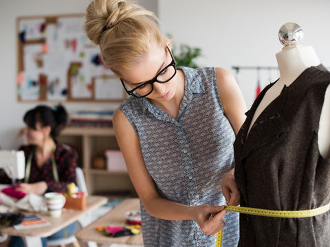 Jeune couturière blonde mesurant une robe noire sur un mannequin dans un atelier, une autre femme floue coud en arrière-plan.