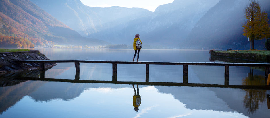 Person in yellow jacket stands on a wooden pier over a calm lake, surrounded by misty mountains. Reflection visible in the water. Serene mood.