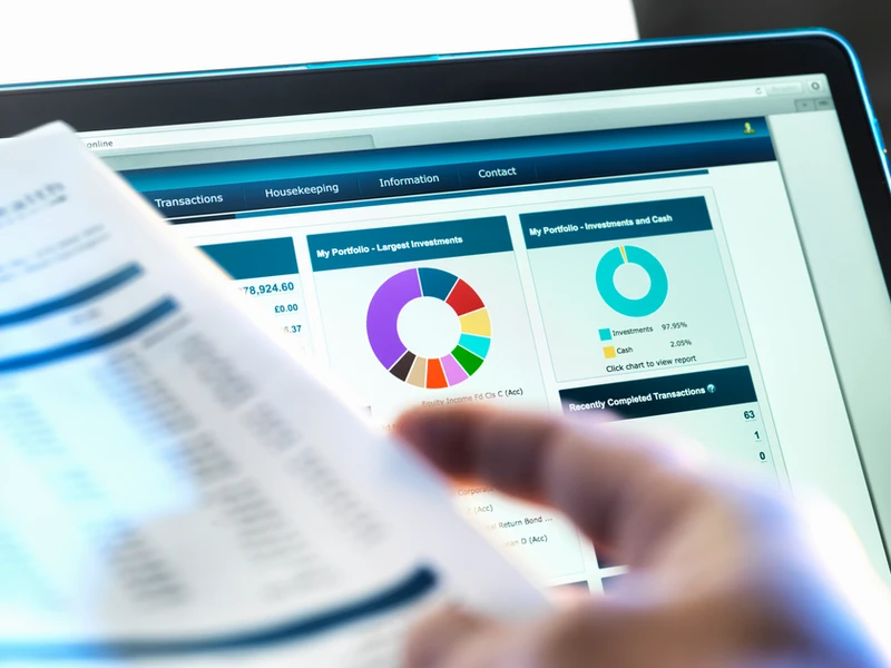 Hand holding documents with charts displayed on a laptop screen. AIM Cube.