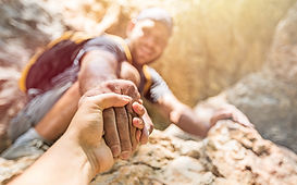 Foreground shows a young Black man bracing on a rocky ledge and extending his hand to pull a White man up from below; both wear outdoor clothing, faces focused, hands locked in a secure grip, and warm sunlight illuminates the textured rock. Composition conveys trust, allyship, crisis intervention, teamwork, and hope, used on a 24/7 mental health resources page to symbolize timely assistance, peer support, and compassionate rescue for people in emotional distress.