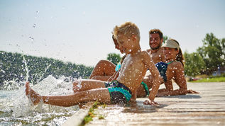 Smiling children in swimsuits splash feet off a sunny lakeside dock, with water droplets spraying and trees in the background
