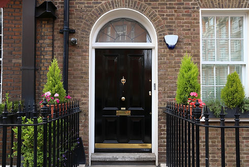 A classic black wooden door with an arch window and brass knocker, framed by brick walls. Potted shrubs and flowers sit beside sleek black railings