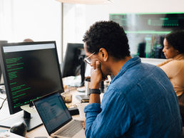 Foreground: Man staring a computer screen with a laptop beneath it. Background: lady working at her desk with other team members computers visible in picture.