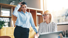 Two businesswomen discuss work in a bright office, one standing with pen and glasses, the other seated at a laptop.