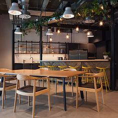 Modern industrial kitchen with wooden tables and chairs, yellow barstools, overhead greenery, and exposed ceiling