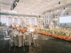 Large reception hall decorated with pink and cream flowers, set tables, an illuminated stage, and a white screen.