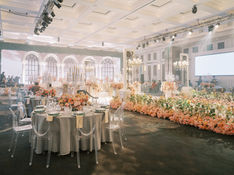 Large reception hall decorated with pink and cream flowers, set tables, an illuminated stage, and a white screen.