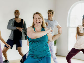 Group dancing joyfully in a bright studio, wearing colorful workout clothes, smiling and energetic. Light streaming through windows.