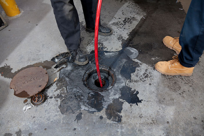 Two workers stand around an open manhole, with a red cleaning hose inserted