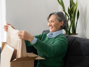 Smiling man sits in a sunlit room at a wooden table with books and photos. Large window, green view outside; casual, relaxed mood.