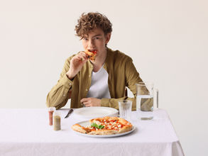 A person in a green shirt enjoys a slice of pizza at a table with a white cloth. A water pitcher and spices are nearby. Bright, neutral backdrop.