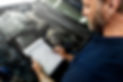 A mechanic with a beard inspects a car engine, holding a checklist on a clipboard