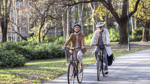 Deux seniors souriants à vélo sur une allée de parc ensoleillée, sous des arbres aux feuilles d’automne.