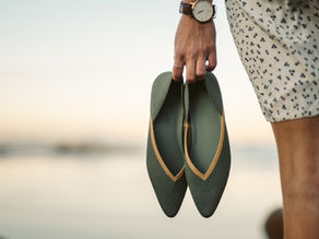 Hand holding green shoes with tan trim by the beach. Person wears a watch and a white dress with black patterns. Soft sunset background.