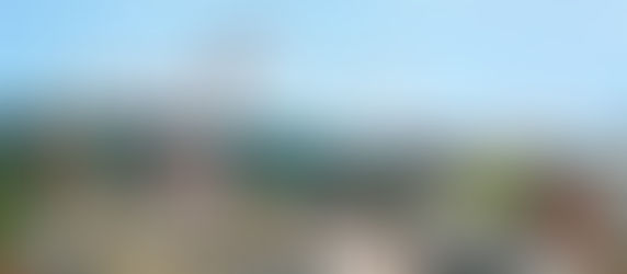 Rooftops of a small town with red cranes towering in the background, set against a backdrop of green hills and a blue sky.