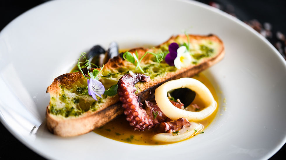 Gourmet seafood plate with octopus, squid rings, pesto toast, and edible flowers in a white bowl on a dark background