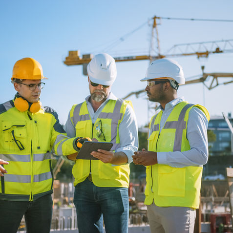 Four construction workers in safety gear converse at a building site with cranes in the background. One holds a tablet, discussing plans.
