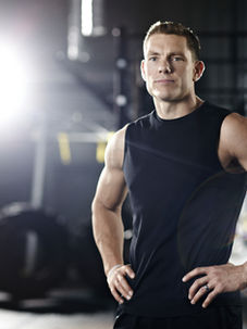 Man in a black sleeveless shirt stands confidently in a gym with tire and equipment in background, illuminated by a bright light.
