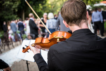 Photo taken from the back of a violinist as he plays as guests congregate.