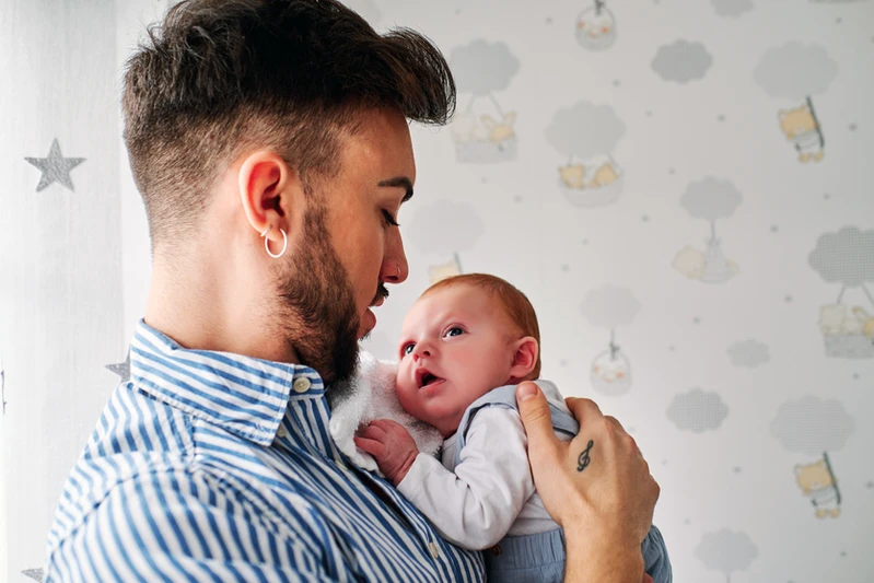 Pai jovem, branco, de camisa azul listrada segurando um bebê no colo.