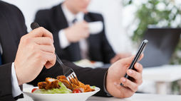 Businessman in a suit eats salad and fried food while checking a smartphone in a bright office meeting room.