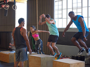 Four people in a gym jump onto wooden boxes. The setting is bright with large windows. They wear athletic gear in vibrant colors.