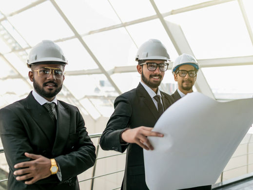 Three men in suits and hard hats review blueprints in a bright, modern building with a glass ceiling. They appear focused and professional.