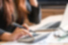A person uses a calculator at a desk with a laptop, papers, and glasses. 