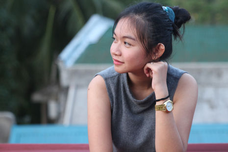 An Asian woman in a grey top gazes thoughtfully to the right, resting chin on hand. Background shows blurred greenery and a building. Wearing a gold watch.