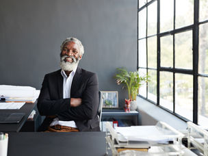 Smiling man in a suit sits at a desk with crossed arms. Sunlit office with large windows, gray wall, plants, and papers scattered around.