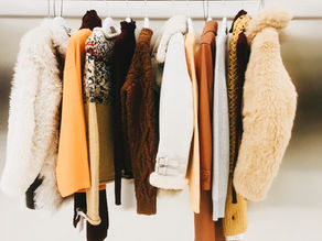 Assorted winter coats and sweaters hang on a rack against a white wall, in neutral, orange, brown, and cream tones.