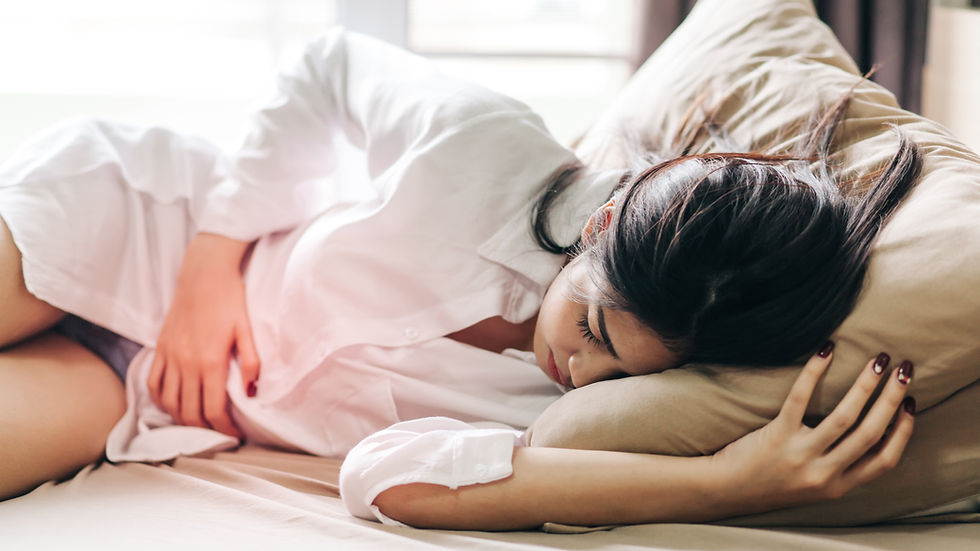 Woman in a white shirt lying on a bed, clutching her abdomen, appearing in pain and discomfort. Reflects women's experience of chronic pain.