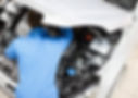 A mechanic in a blue uniform works under the hood of a white car, focusing on the engine