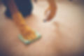 A person in yellow gloves cleans a beige carpet with a spray bottle and cloth