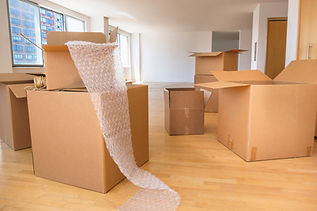 An empty room with large windows and a wooden floor, filled with open cardboard boxes and bubble wrap