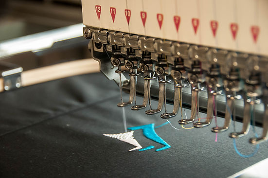 An embroidery machine stitches "HTH" in gold on navy fabric