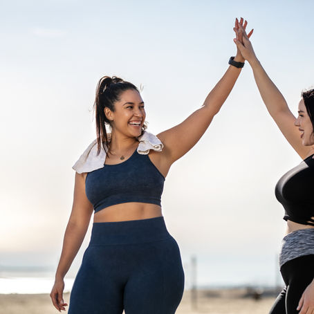 Two women in workout clothes high-five on a sunny beach. One wears a towel around her neck. They are smiling, exuding a joyful mood.