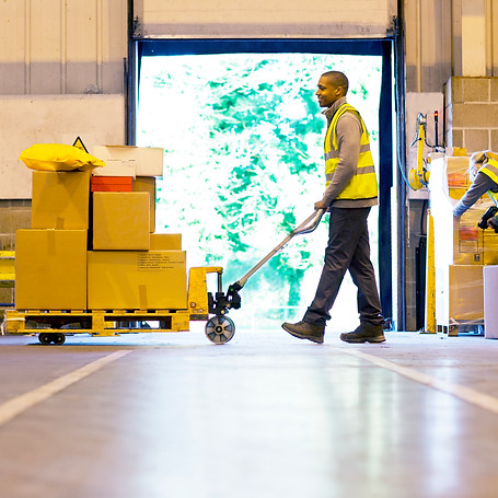 Worker in hi-vis vest pulling a hand truck loaded with boxes in a warehouse.