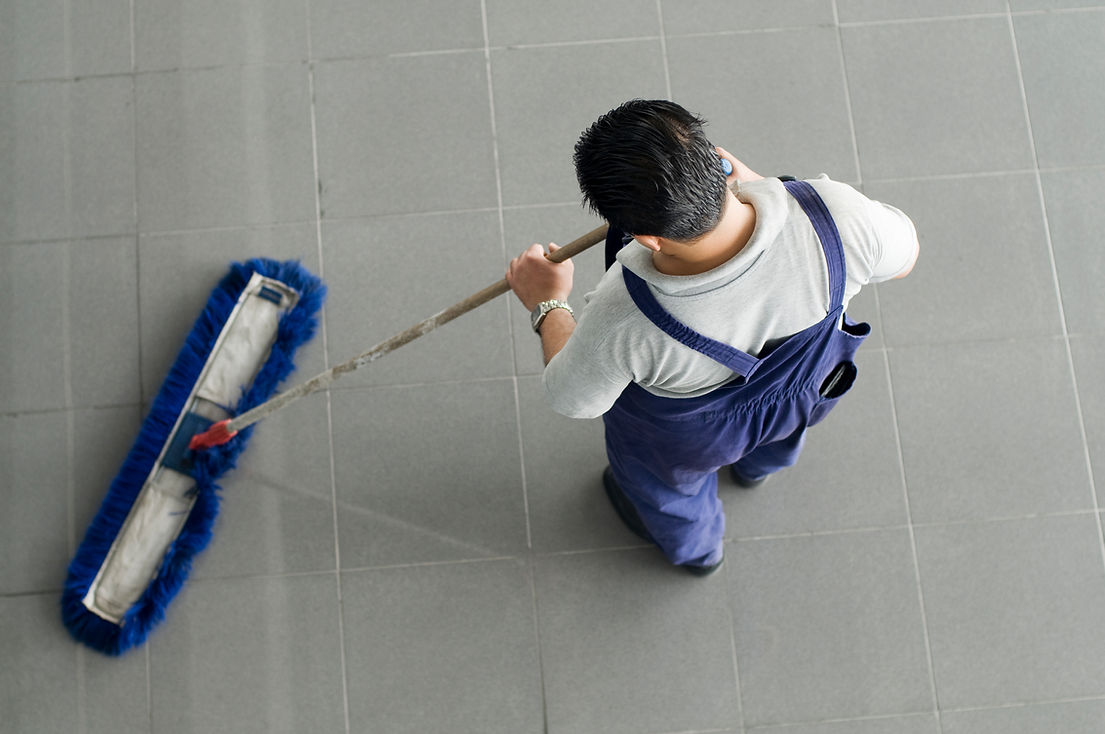 A person in blue overalls sweeps a large gray tiled floor with a wide, blue-bristled broom