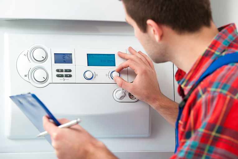 A person in a plaid shirt and blue overalls adjusts a thermostat on a white heating system