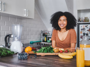 A woman smiling in her kitchen, surrounded by fresh fruits, greens, and a blender, ready to make a healthy smoothie.